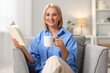 © New Africa - Smiling middle aged woman with cup of hot drink reading book at home
