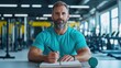 © Pixelbiz - New Year's resolutions personal goals skills concept. A focused man in a gym writes in a notebook, showcasing fitness dedication and planning, with weights visible in the background.