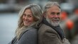 © liliyabatyrova - A joyful mature couple sits back to back on a cool autumn day by the waterfront, embracing the moment with smiles that reflect their love and companionship as they enjoy the serene atmosphere
