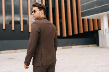 Wall Mural - Rear view of photogenic confident businessman in sunglasses and brown suit standing posing outdoors on summer day. Handsome bearded business man exuding confidence and determination.