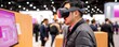 © Ramita - Consumer Electronics Show concept. A person experiences virtual reality technology at an exhibition, wearing a VR headset and interacting with a display in a bustling environment.