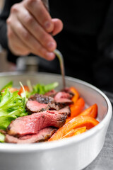 Wall Mural - A close-up of a chef's hand plating a colorful salad with sliced roast beef, fresh greens, and vibrant red peppers, highlighting culinary artistry and delicious presentation.