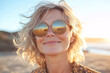 © youriy - A cheerful blonde woman with wavy hair wearing round sunglasses reflecting the beach at sunset radiating warmth and happiness in natural light
