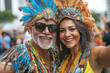© Helena GARCIA - Pareja senior brasileña, de hombre y mujer de mas de 50 años disfrazados con plumas y trajes coloridos en el carnaval de Rio en Brasil con fondo desenfocado de una calle urbana
