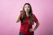 © Daniel - Asian woman in a red t-shirt and jeans standing against a pink background, smiling brightly with excitement, raising one finger upward as if having a new idea or solution
