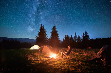 Wall Mural - Hiker woman relaxes by campfire, illuminated by its warm glow, with tent nearby under stunning, star-filled sky. Milky Way stretches overhead, adding magical touch to serene mountain setting.