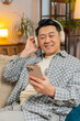 © Andrii Iemelianenko - Happy overjoyed middle-aged man in wireless headphones relaxing sitting on couch using phone at home choosing, listening favorite energetic disco dancing music. People weekend leisure activities.