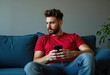 © Vibudhaart - A young man with a beard sits on a couch and looks at his phone.