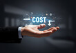 © Shanewaz - Business concept of a hand holding the word 'COST' with financial data icons and a downward arrow, symbolizing cost reduction, financial management, and economic efficiency.