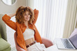 © SHOTPRIME STUDIO - Young woman with curly hair wearing casual outfit, smiling while working on laptop at home Cozy workspace with warm colors and natural light