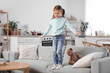 © Pixel-Shot - Cute little girl standing on edge of sofa at home. Child in danger