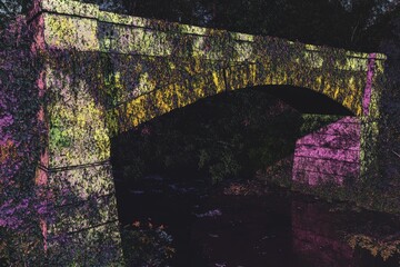 Sticker - A vibrant, overgrown stone bridge arching over a tranquil stream, surrounded by nature.