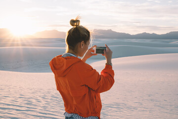 Wall Mural - back view of young woman using smartphone camera for making picture of sunset in white sand dunes at