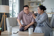 © ultramansk - An elderly couple sits in their living room. The man holds his knee, appearing to be in discomfort, while his partner offers support. The image conveys care, concern, and the bond between the couple.