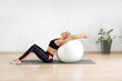 © junky_jess - Fit adult woman in black sportswear sits on a mat and does back stretching using fitness ball in studio indoor, side view.