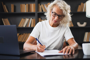 Middle aged senior woman using laptop computer writing notes at workplace. Focused mature old businesswoman work at office making written records doing online work. Mature boss professional worker