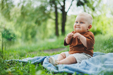 Wall Mural - Little boy 1 year old sitting on the grass in the park and eating bread. Baby on a walk in the fresh air.