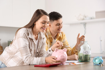 Wall Mural - Young couple putting coin into piggy bank at table in kitchen