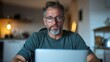 © Dulemegapixel - A contemplative man with glasses and grey hair sits quietly, working on his laptop in a serene home environment, suggesting a peaceful, productive task-focused moment.