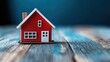 © Dulemegapixel - A striking red model house placed on a blue wooden background, emphasizing bold colors and depicting real estate and property investment themes vibrantly.