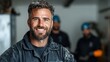 © Dulemegapixel - A cheerful man in a black uniform smiles confidently, with two workers in the background, demonstrating teamwork and dedication in a dynamic industrial setting.