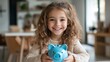 © Dulemegapixel - A cheerful young girl holding a blue piggy bank, dressed in a knitted sweater, is indoors with a cozy background, radiating warmth and innocence.
