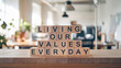 © E - Wooden blocks stacked vertically display 'LIVING OUR VALUES EVERYDAY,' set against a softly blurred office backdrop with natural lighting, conveying a motivational and professional theme.