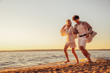 Smiling beautiful woman and her handsome boyfriend. Excited couple in casual clothes. Happy cheerful family. Female and man run at sunrise over sea beach outdoors. Run at seaside in summer day