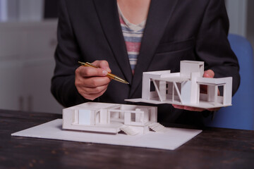 Wall Mural - A businessman sits at a table at night, holding a model of a two-story white house. He reviews designs, drafts plans, and inspects the roof details, showcasing modern architecture