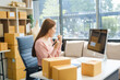 © NanSan - An Asian businesswoman sits at her desk, working on online sales and e-commerce marketing strategies. As a small business owner, she prepares and packages product boxes to ship to customers.
