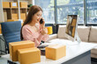 © NanSan - An Asian businesswoman sits at her desk, working on online sales and e-commerce marketing strategies. As a small business owner, she prepares and packages product boxes to ship to customers.