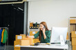 © NanSan - An Asian businesswoman sits at her desk, working on online sales and e-commerce marketing strategies. As a small business owner, she prepares and packages product boxes to ship to customers.