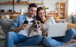 © Nikhil - A happy young couple sitting together on a comfortable sofa at home, using a laptop and mobile phone while hugging their dog, representing the online shopping concept