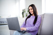 © deagreez - Charming young woman enjoying leisure in stylish casual wear, smiling while using laptop sitting on a cozy couch, filled with daylight