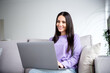 © deagreez - Young woman with brunette hair smiling while using a laptop in a cozy living room, enjoying leisure time at home