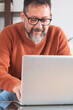 © simona - Happy smiling middle aged older mature man wearing glasses looking at laptop using computer writing notes watching webinar sitting at home table. Hybrid work, elearning, online learning concept.