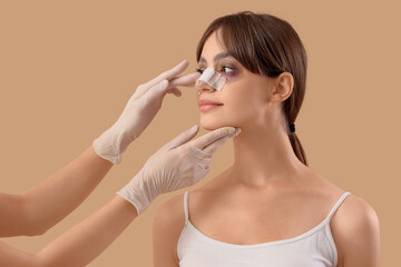 Surgeon hands touching woman's bandaged nose after plastic surgery on beige background