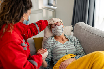 Wall Mural - Female emergency responder in red uniform monitors elderly patient wearing protective masks during home healthcare visit. Patient rests comfortably on gray couch.