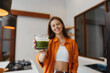 © SHOTPRIME STUDIO - Smiling young woman holding a green smoothie in a cozy kitchen, promoting healthy eating and wellness, vibrant orange outfit and natural light