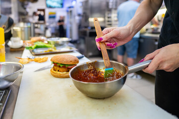 Wall Mural - A chef combines ingredients in a stainless steel bowl, preparing sauce in a bustling kitchen. A freshly made burger sits nearby, highlighting a vibrant culinary scene.