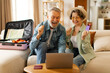 © Home-stock - Happy senior couple booking flight online with laptop and credit card, sitting with packed suitcase on sofa at home