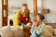 © Home-stock - Loving senior man giving bouquet of flowers and gift box to surprised wife, sitting on sofa at home. Birthday, anniversary or Valentine's day celebration