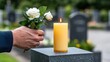 © lililia - Hands carefully place a yellow rose next to an incense candle on a tombstone, with a man in black mourning nearby surrounded by white roses