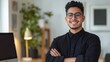 © ifoto - A happy young Latin man with a black shirt, casual outfit, and glasses is standing in his modern office space with his arms crossed, smiling at the camera.