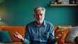 © Pete - Senior man sits on orange couch in living room at night. Looks confused, uncertain. Feeling embarrassed frustrated by ambiguous question. Arms open as searching for answer. Indoor setting shows teal