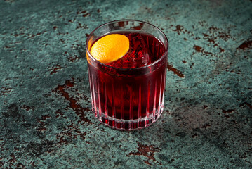 Wall Mural - A refreshing cocktail with a deep red color, served in a stylish glass with ice and a vibrant orange slice garnish, set against a textured, rustic background. Perfect for bar or drink-themed designs.