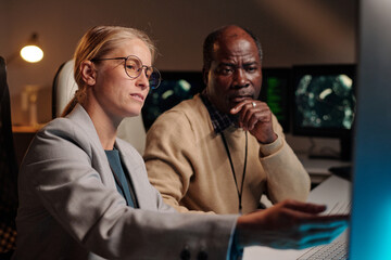 Young confident blond female IT engineer pointing at computer screen while explaining male colleague how to decode data