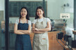 © Wicitr - Smiling Cafe Owners: Two young Asian women, cafe owners, stand proudly with arms crossed in front of their open shop, radiating confidence and welcoming customers.
