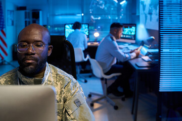 Aufkleber - Serious young man in camouflage uniform sitting in front of desktop computer against coworkers in military office