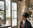 © larrui - Pensive woman in a rustic kitchen, sitting by a sunlit window with stone walls.